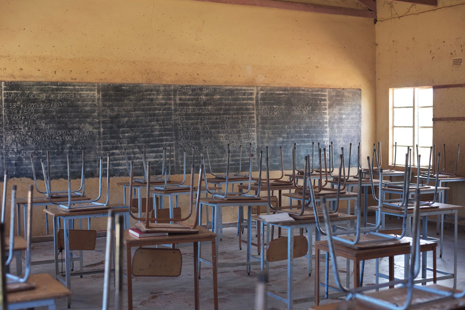 An empty classroom featuring wooden desks with chairs set upside down on top and a blackboard covered in chalk writing.
