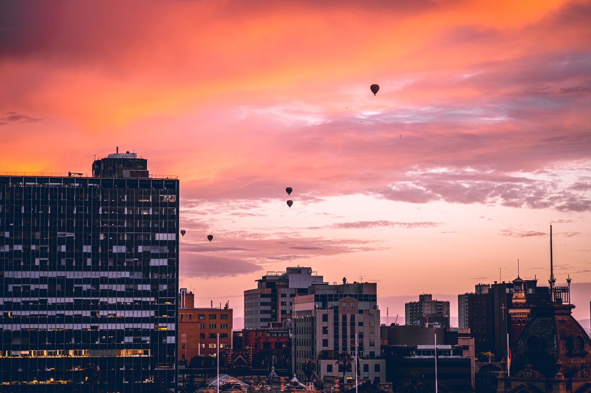 Hot air balloons drifting in a pink and orange sunset sky above a city skyline with an office tower and older buildings.