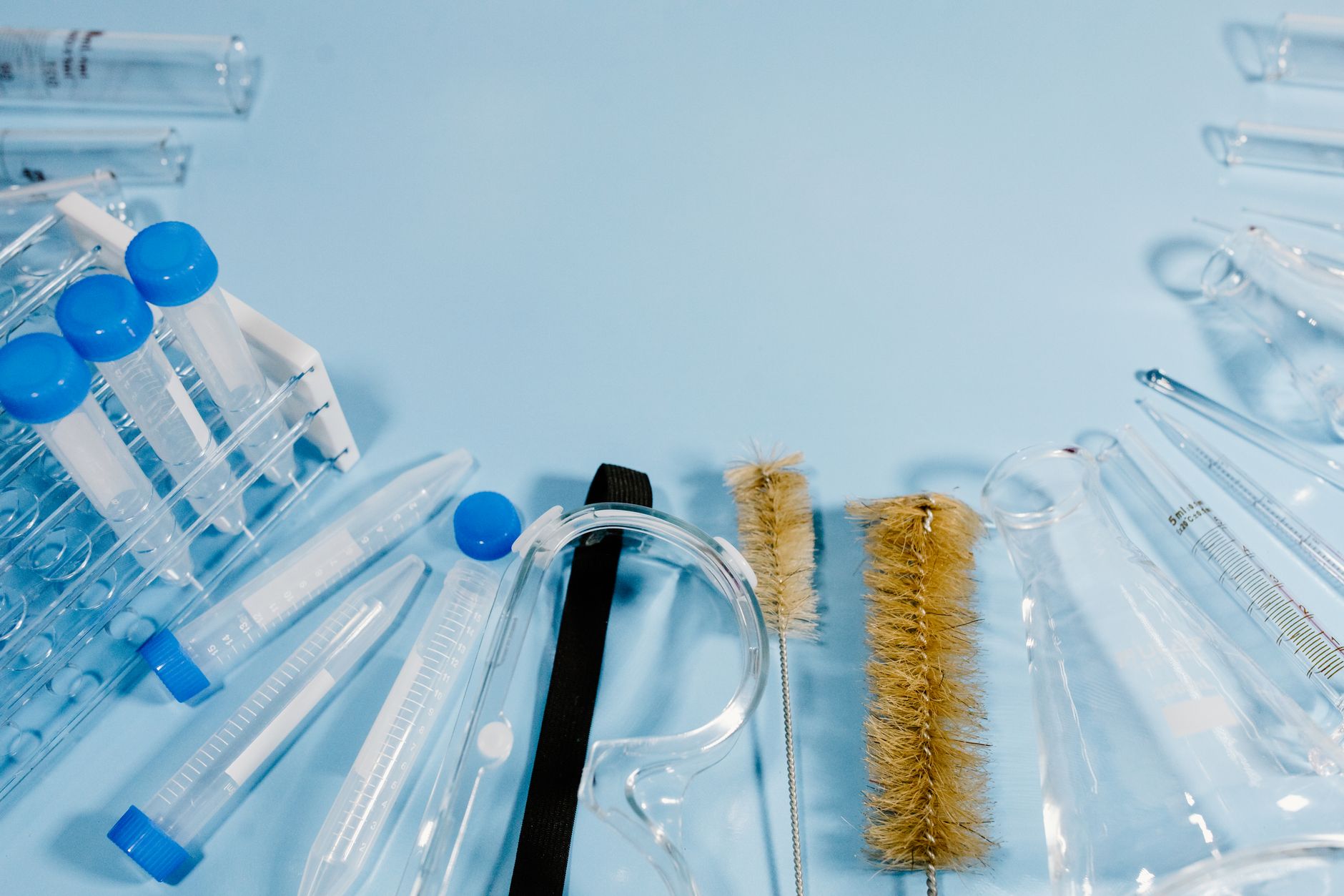 Laboratory glassware, plastic centrifuge tubes, test tube brushes, and safety goggles arranged on a blue surface.