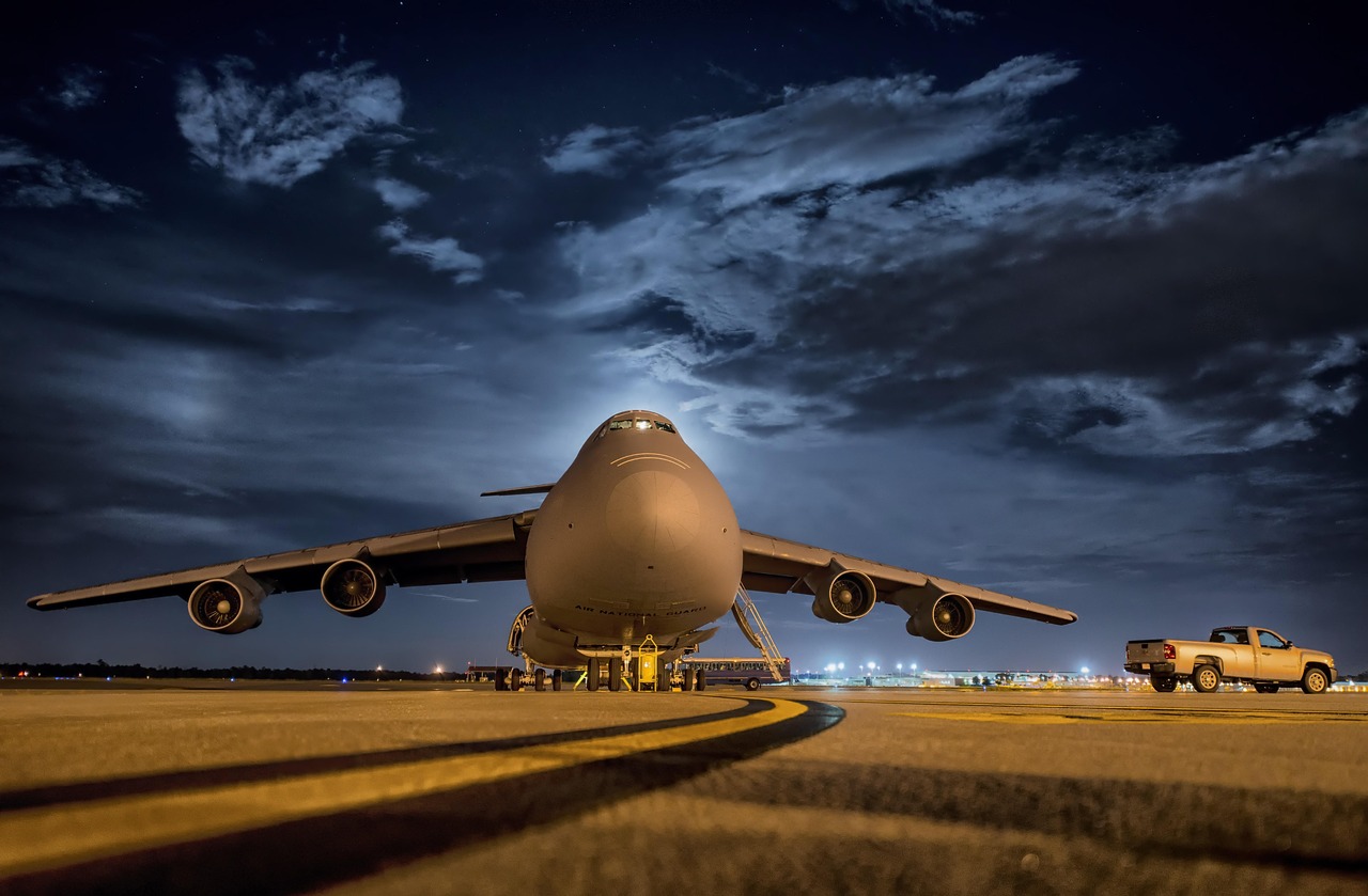 Large military transport aircraft parked on a floodlit apron at night, a pickup truck beside it under a stormy sky