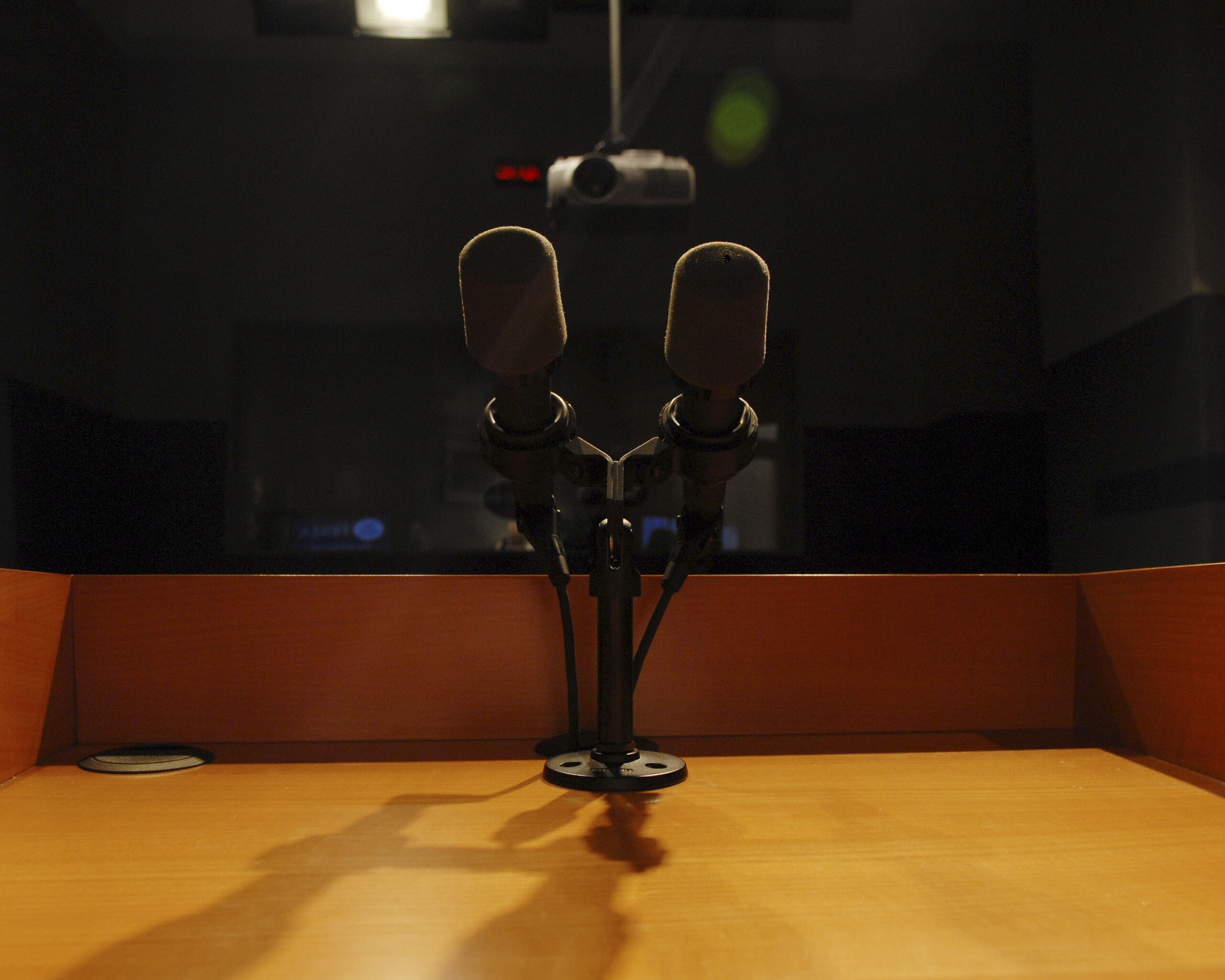 Two microphones with foam windscreens on a stand atop a wooden podium, in a dark room with a projector hanging from the ceiling.