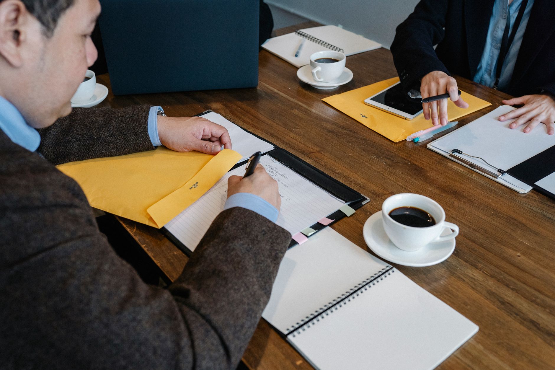 Two people at a wooden table write notes on papers and a clipboard, with yellow envelopes, a tablet, notebooks and cups of coffee around them.