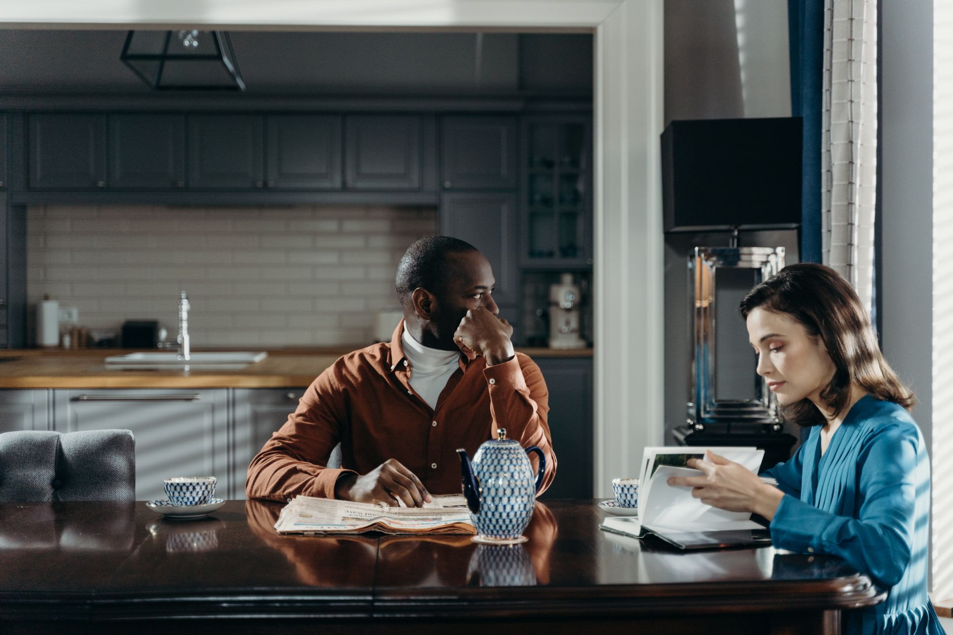 Un homme et une femme à une table de cuisine: il a un journal ouvert devant lui, elle feuillette un livre, avec une théière et des tasses sur la table.
