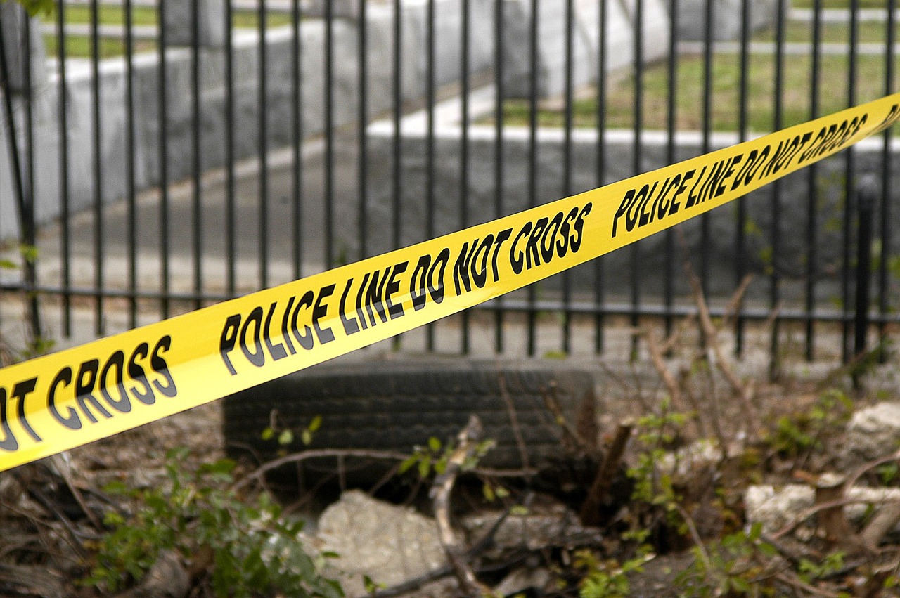 Yellow police tape printed with a warning stretched across a black metal fence, with a discarded tyre lying in weeds below.
